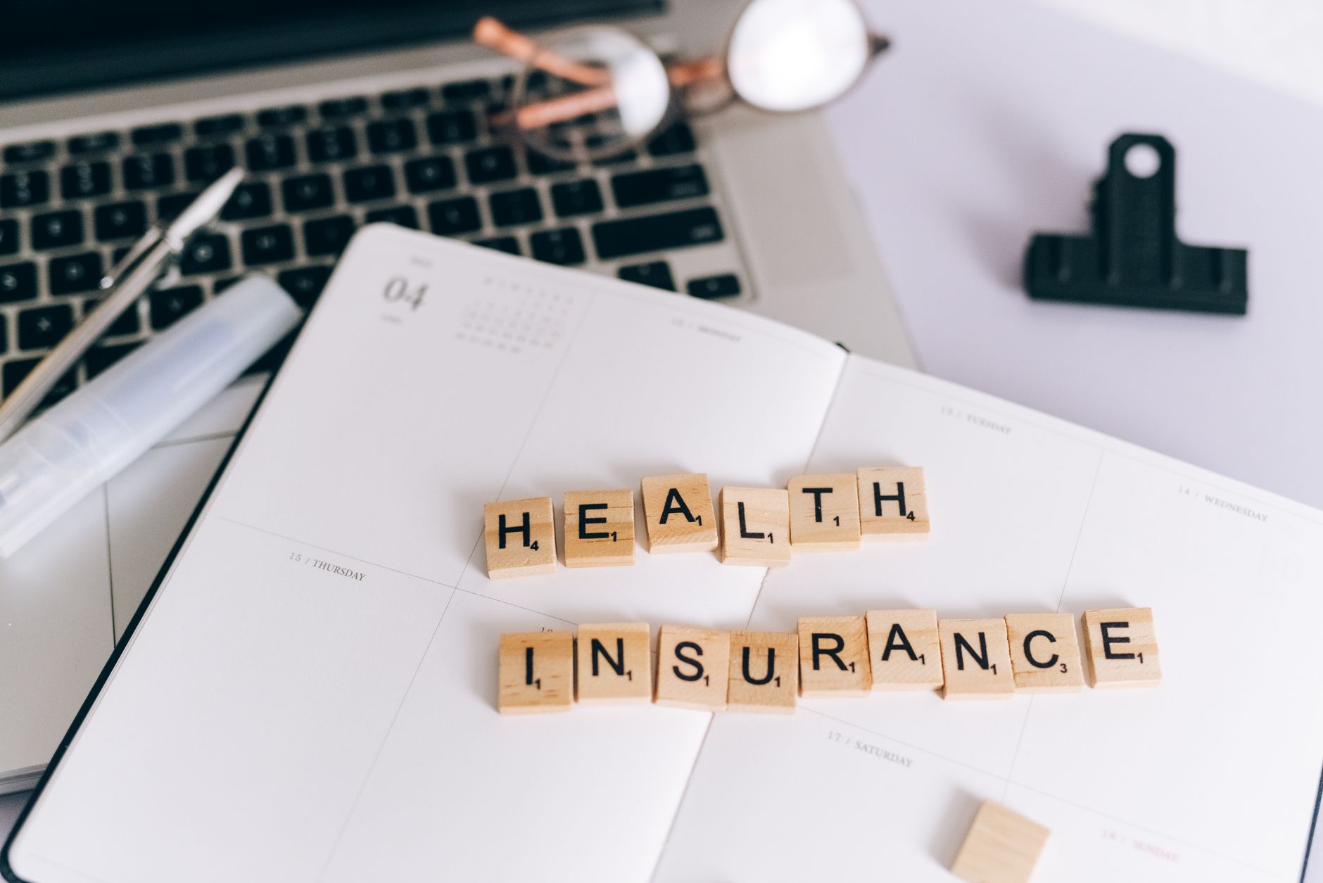 Close-up of Scrabble tiles spelling 'Health Insurance' on a planner with a laptop. — Bruce Hensel: Promoting Healthy Choices