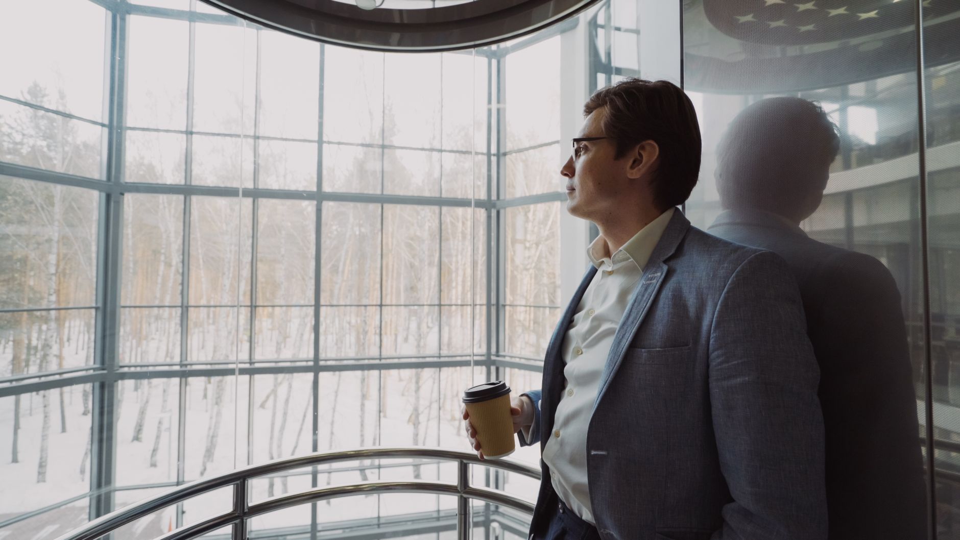 Businessman in blue suit with coffee cup looking out a large window, reflecting on a winter landscape. — Designing Perfection