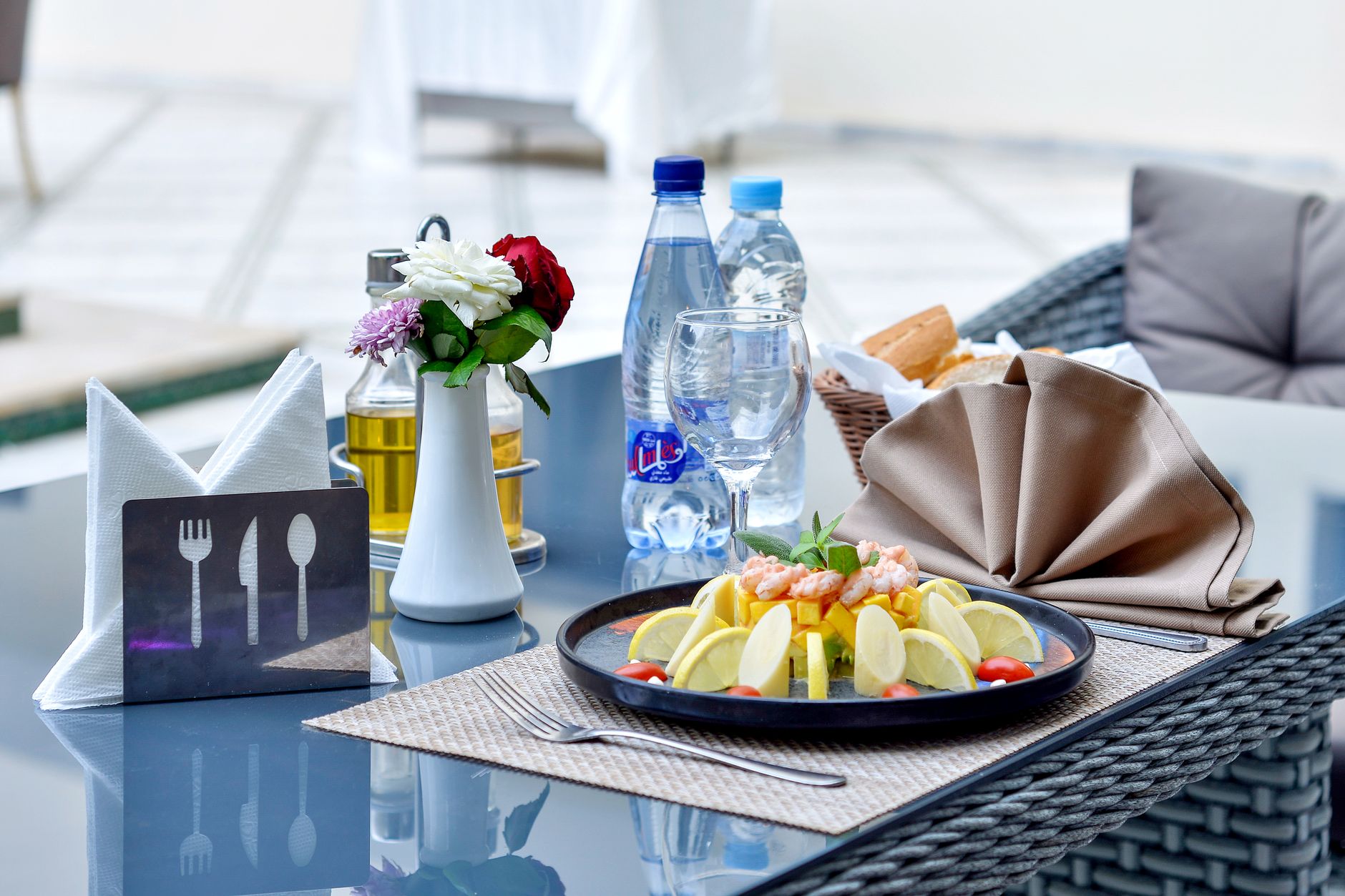 A stylish outdoor restaurant table featuring a seafood dish with lemon slices, bottled water, and decorative cutlery. — Why M